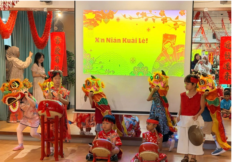 Children perform a lion dance and drum in front of a screen reading Xin Nian Kuai Le! with red decor